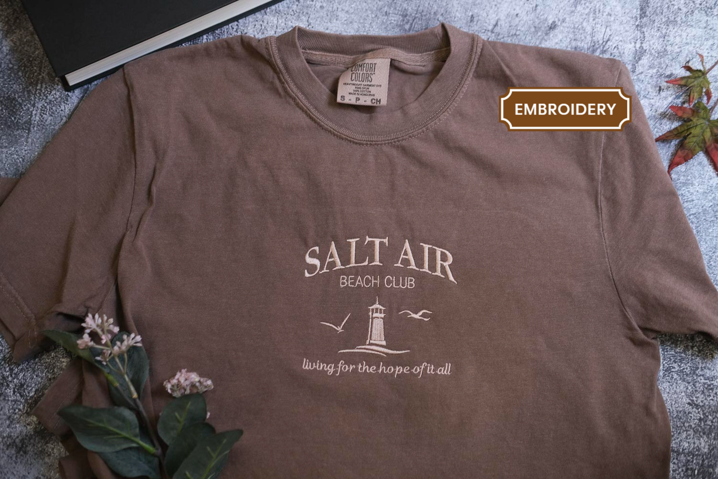 A brown t-shirt with the text "SALT AIR BEACH CLUB" printed on it, along with a lighthouse and birds, is displayed on a gray surface.