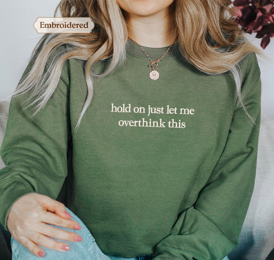 A person wearing a green sweater with the text "hold on just let me overthink this" embroidered on it.