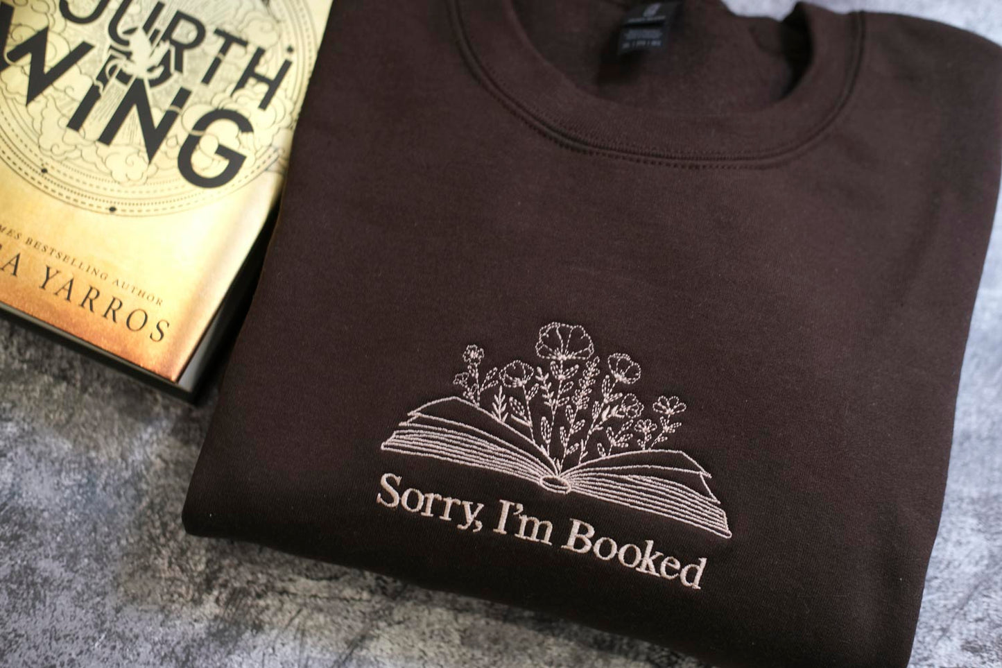 A dark-colored sweatshirt with a white embroidered design on the front, featuring a book and flowers, and the text "Sorry, I'm Booked" printed on it.