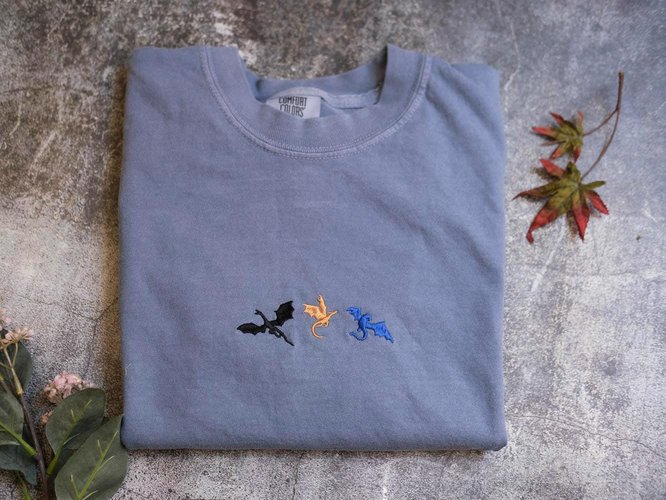 a gray t-shirt with three dragon embroidered designs on it, placed on a gray surface alongside some leaves.