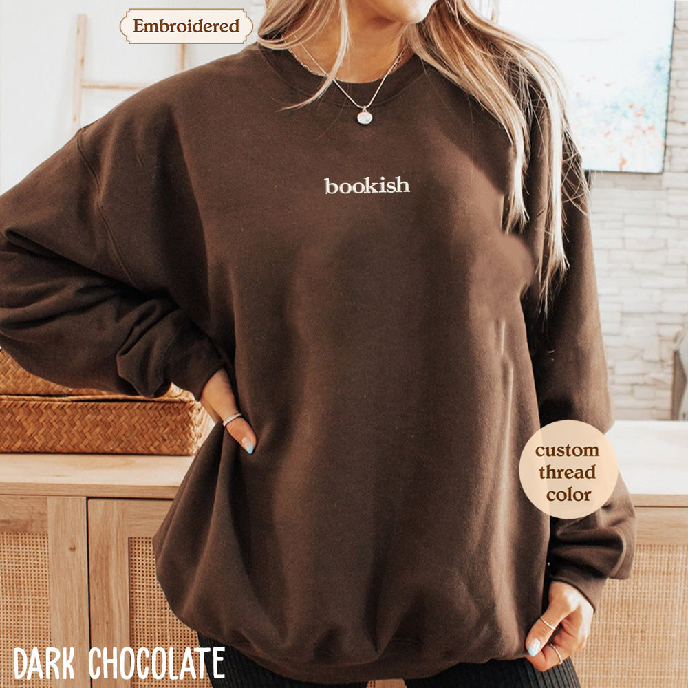 A woman wearing a brown sweater with the word "bookish" printed on it, standing in front of a wooden cabinet.