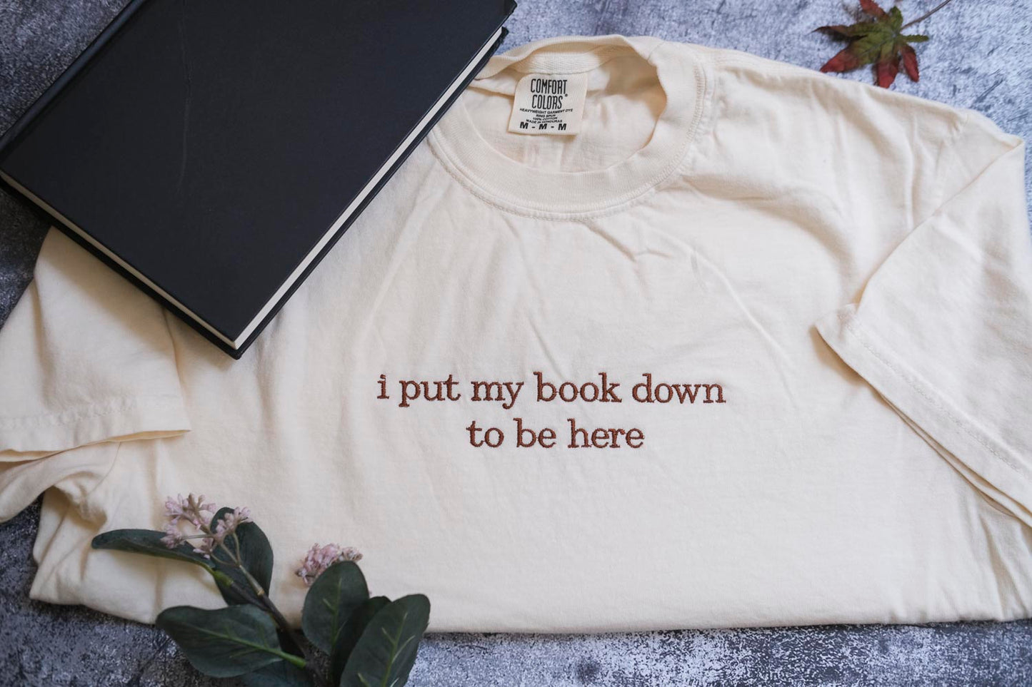 a white t-shirt with the text "I put my book down to be here" printed on it, along with a black book and some flowers.
