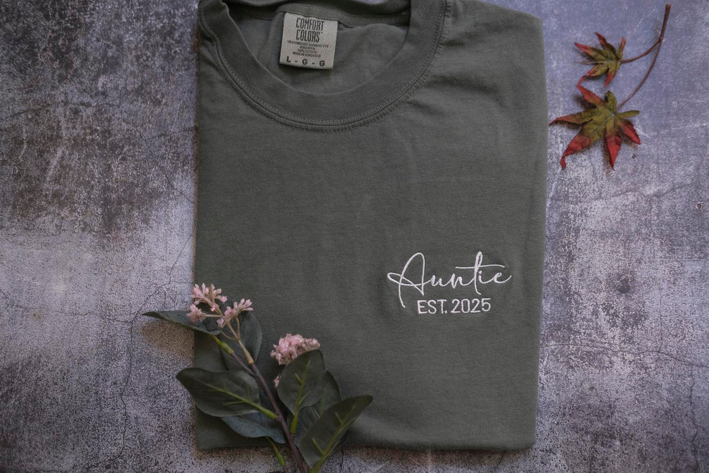 a gray t-shirt with the text "Auntie EST 2025" printed on it, along with some leaves and flowers.