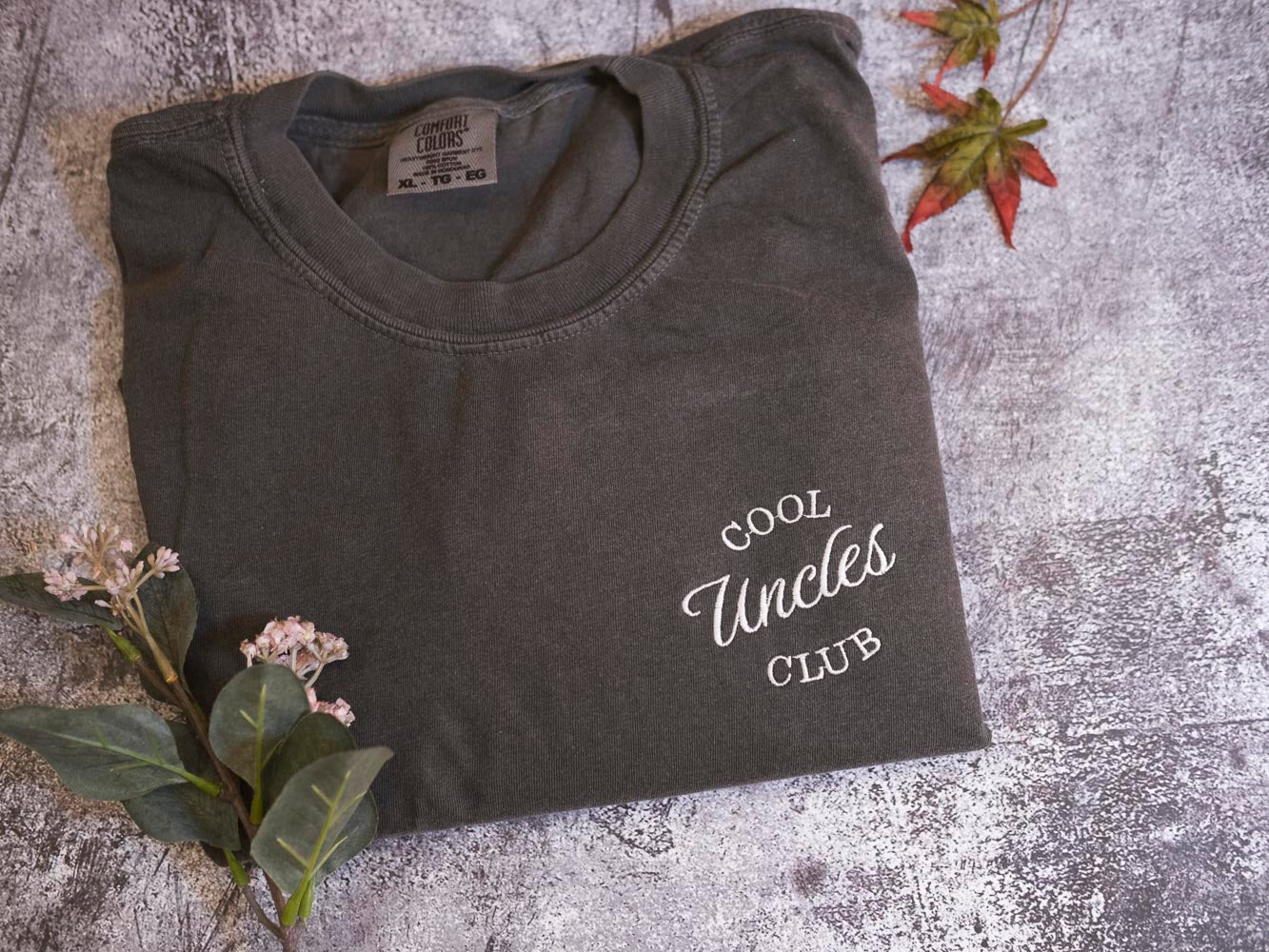 a gray t-shirt with the text "Cool Uncle Club" printed on it, along with some leaves and flowers on a textured surface.