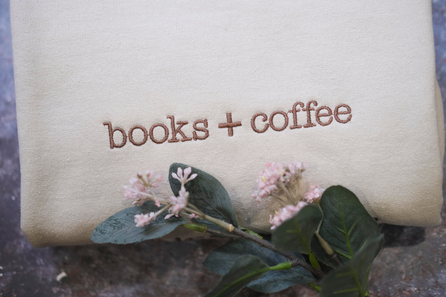 a beige fabric with the text "books + coffee" embroidered on it, along with a small branch of pink flowers and green leaves.