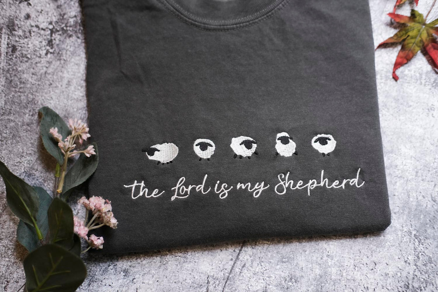 A black t-shirt with a white sheep design and the text "The Lord is my Shepherd" printed on it, along with some leaves and flowers.