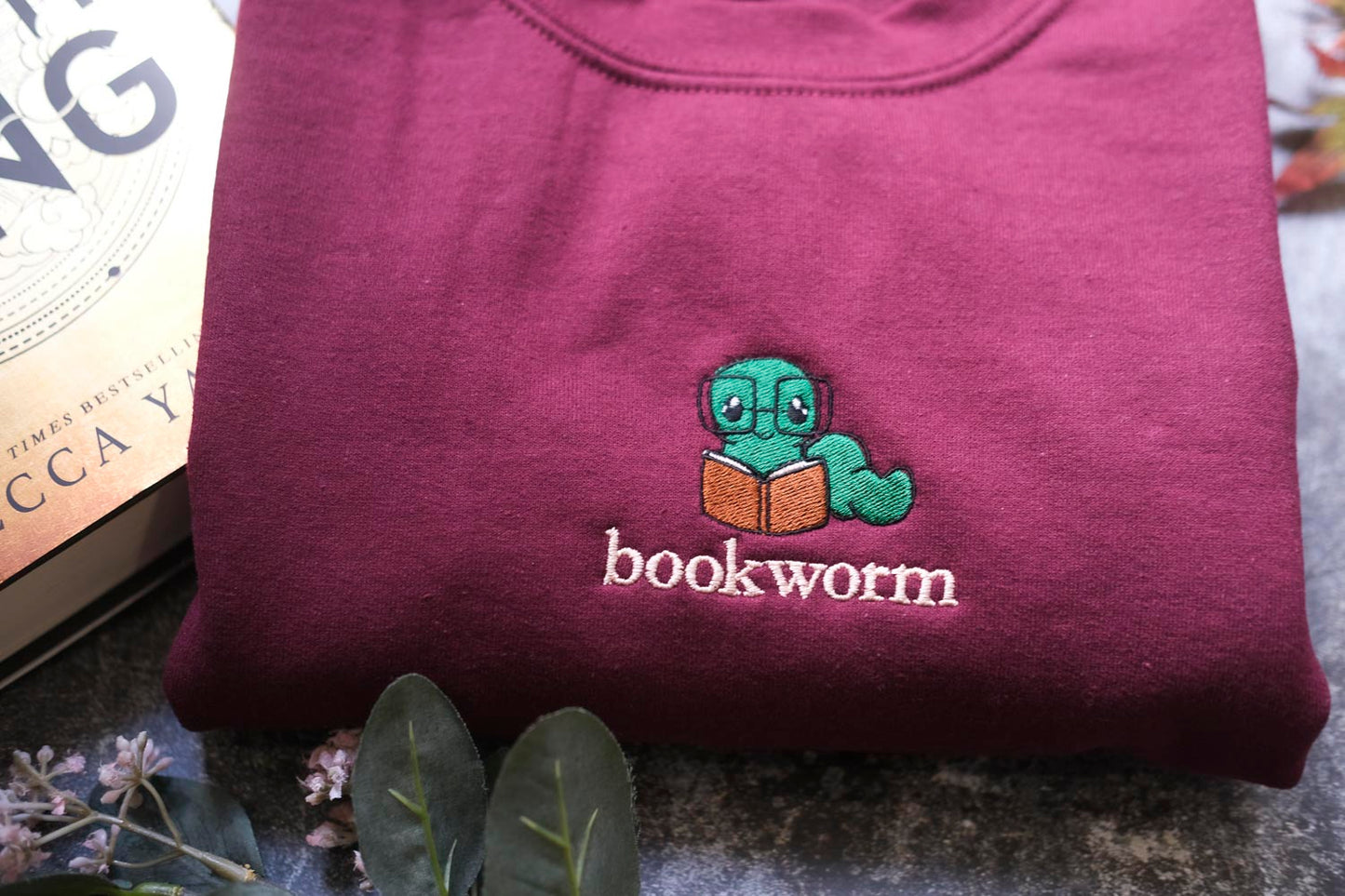 A maroon sweatshirt with a small green book worm embroidered on it, along with a book and some flowers.