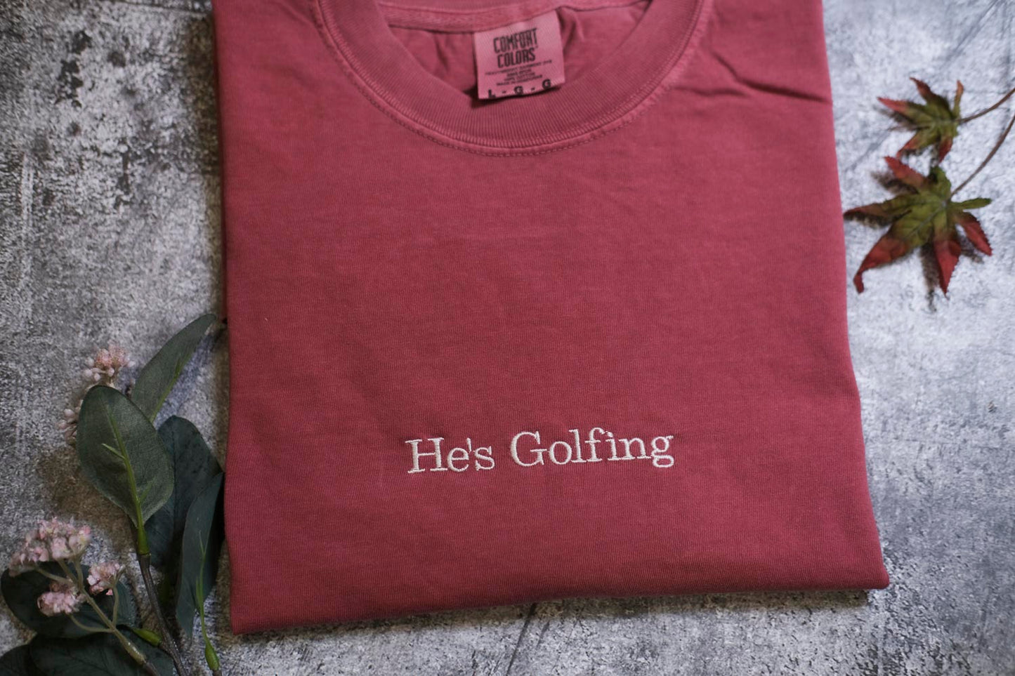A red t-shirt with the text "He's Golfing" printed on it, along with some leaves and flowers.