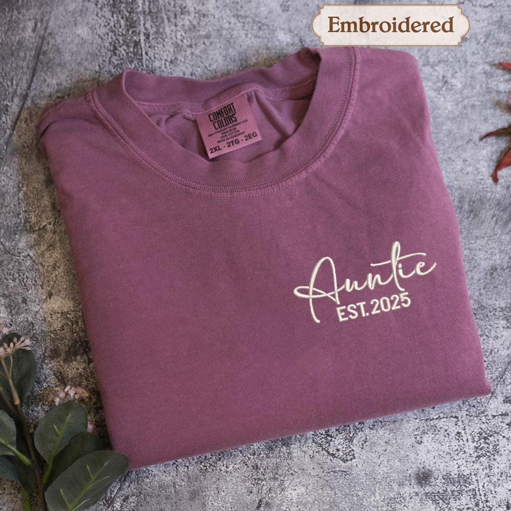 A purple t-shirt with the text "Auntie EST.2025" embroidered on it, along with a label indicating it is "Embroidered" and "Comfy Colors" in the neck area.