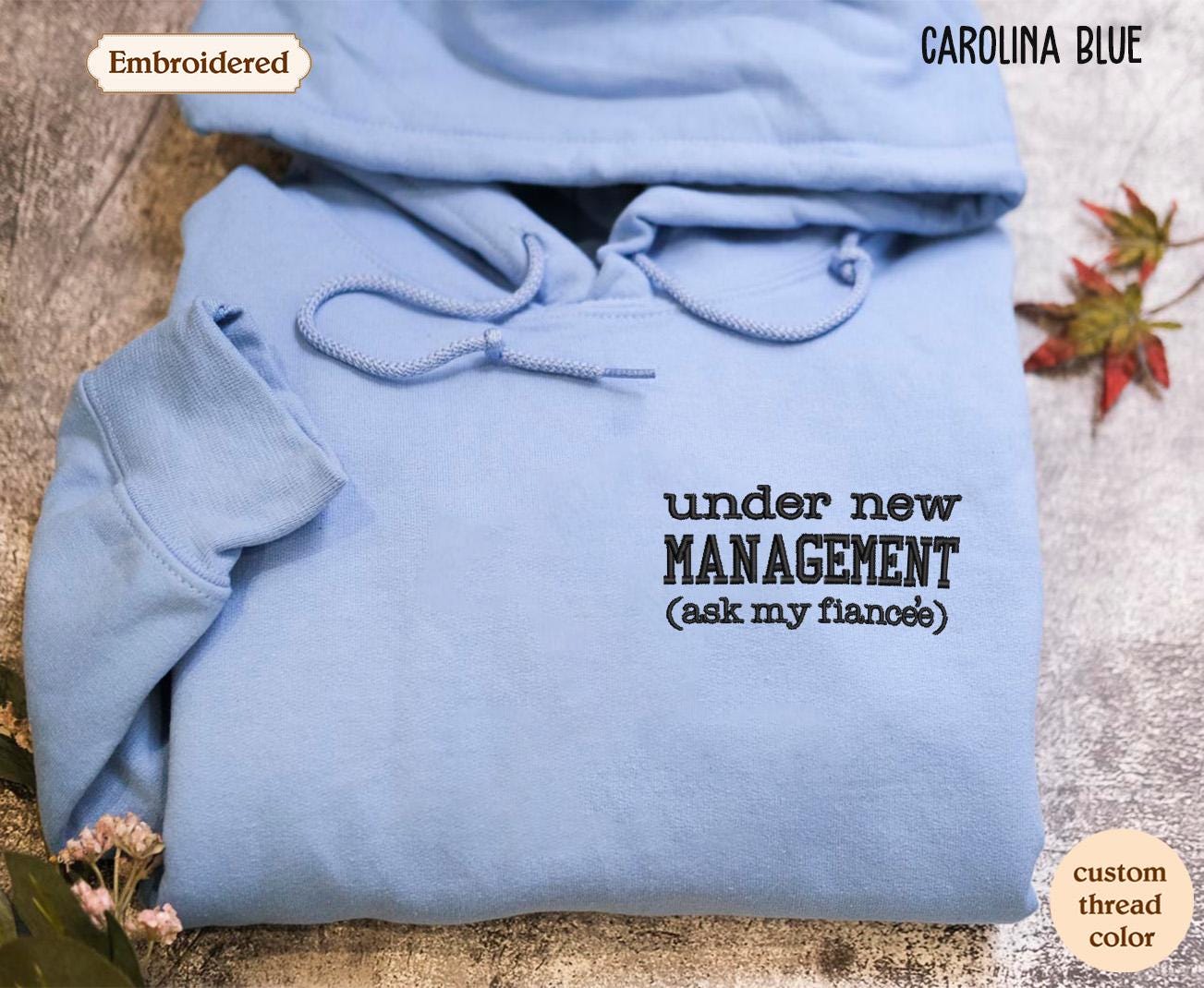 A light blue hoodie with the text "under new MANAGEMENT (ask my fiancee)" printed on it, along with the text "Carolina Blue" at the top.