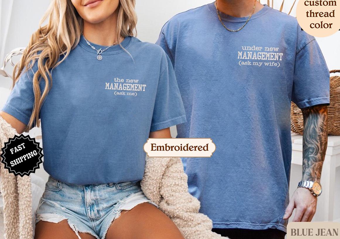A woman wearing a blue t-shirt with the text "the new MANAGEMENT (ask me)" and a man wearing a blue t-shirt with the text "under new MANAGEMENT (ask my wife)" standing next to each other.