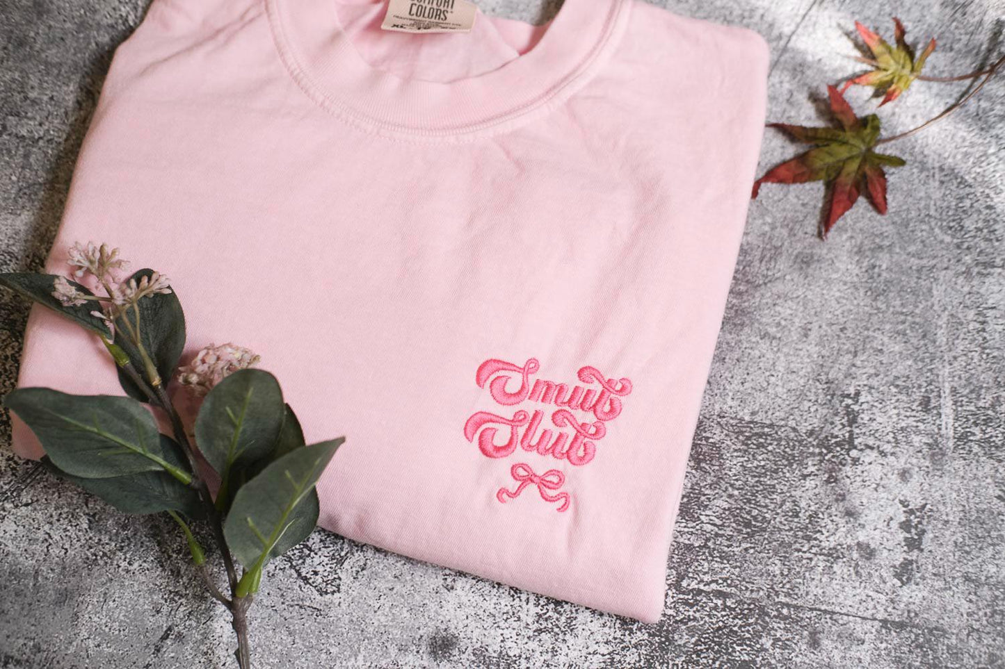 A pink t-shirt with a pink embroidered text on it, along with some leaves and a small flower, placed on a gray surface.