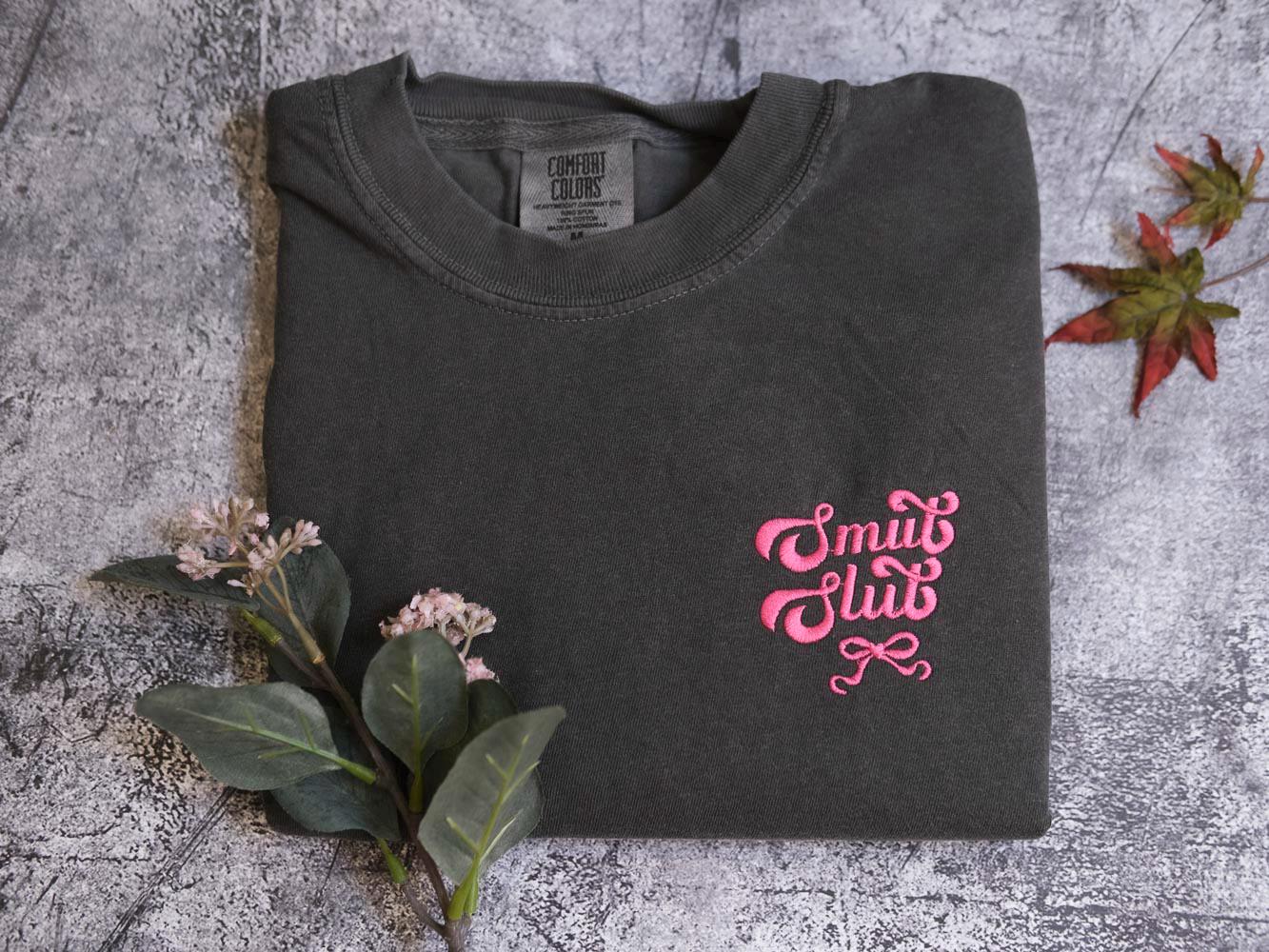 a gray t-shirt with pink text and a small leaf on a textured surface.
