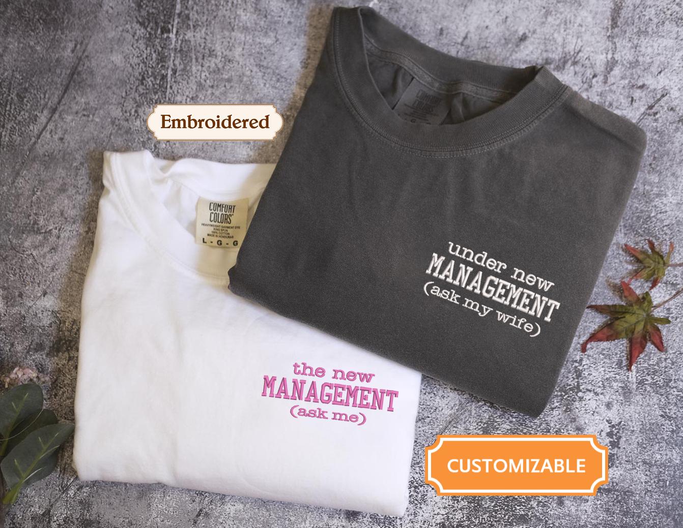 two t-shirts with humorous text printed on them, one white and one gray, placed on a textured surface.
