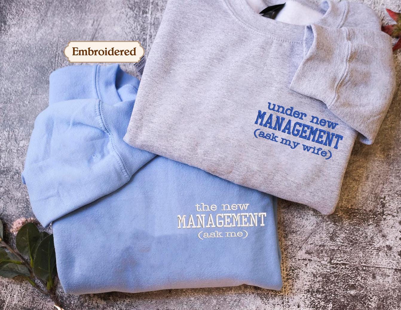 two shirts, one blue and one gray, with embroidered text on them. The blue shirt has the text "Embroidered" and "under new MANAGEMENT (ask my wife)" on it, while the gray shirt has the text "the new MANAGEMENT (ask me)" on it.
