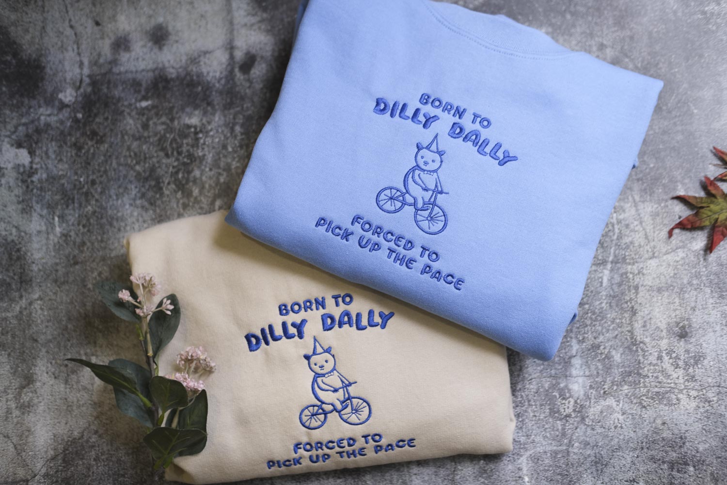 two sweatshirts with a blue and a beige color scheme, each with a unique design and text. The blue sweatshirt has a cartoon image of a cat riding a bicycle and the text "Born to Dilly Dally" and "Forced to Pick Up the Pace". The beige sweatshirt has a similar cartoon image of a cat riding a bicycle and the text "Born to Dilly Dally" and "Forced to Pick Up the Pace".