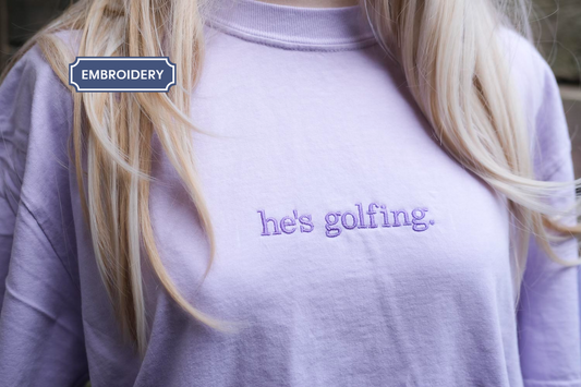 A person wearing a purple t-shirt with the text "he's golfing" printed on it.