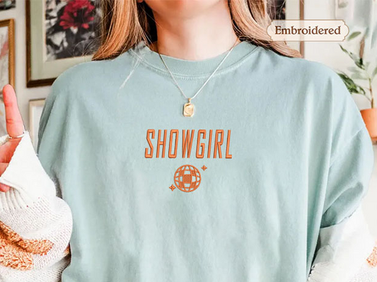 A person wearing a light blue t-shirt with the word "SHOWGIRL" printed on it, along with a small embroidered globe design.