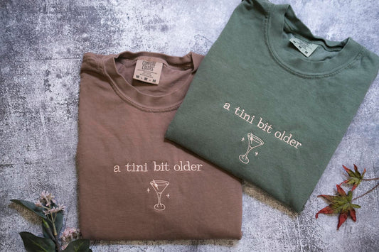 Two t-shirts with the text "a tini bit older" printed on them, along with a cocktail glass icon, are laid out on a textured surface.