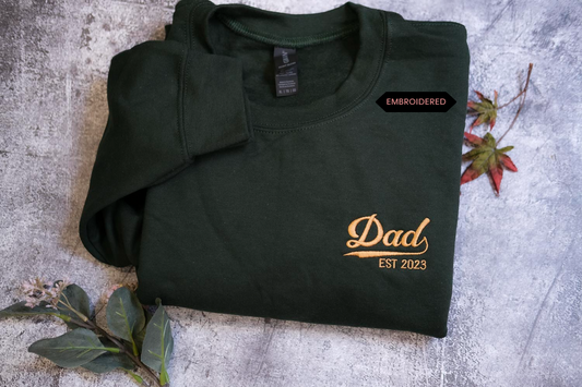 A dark green sweatshirt with the text "DAD EST 2023" printed on it, along with a small gold-colored logo.