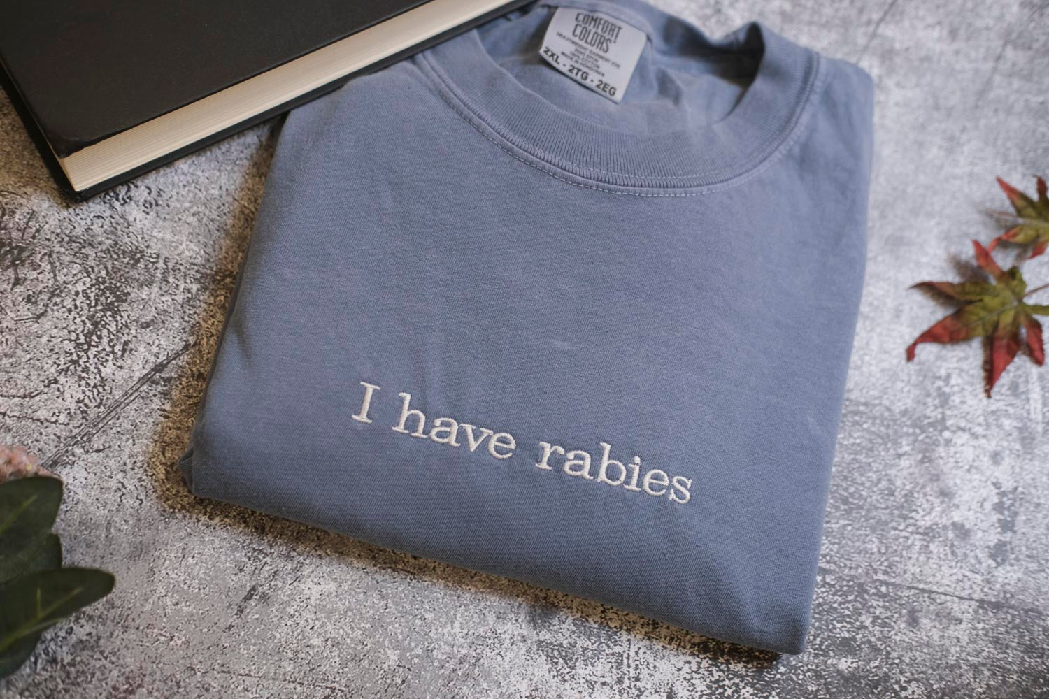 A gray t-shirt with the text "I have Rabies" printed on it, along with a black book and some leaves on a textured surface.