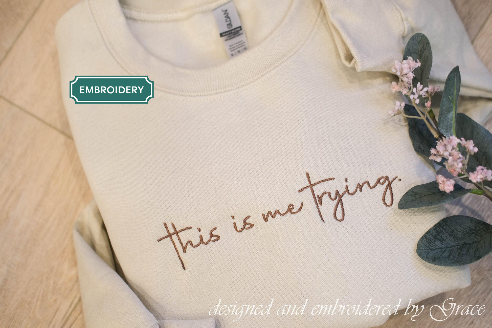 a white t-shirt with the text "This is me trying" embroidered on it, along with a green embroidery label and a small floral arrangement.