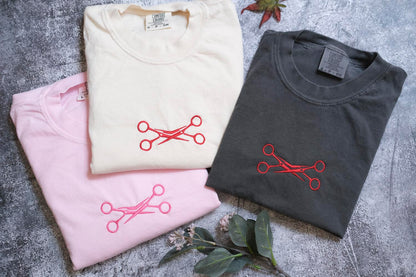 three t-shirts with scissors embroidered on them, arranged on a gray surface.