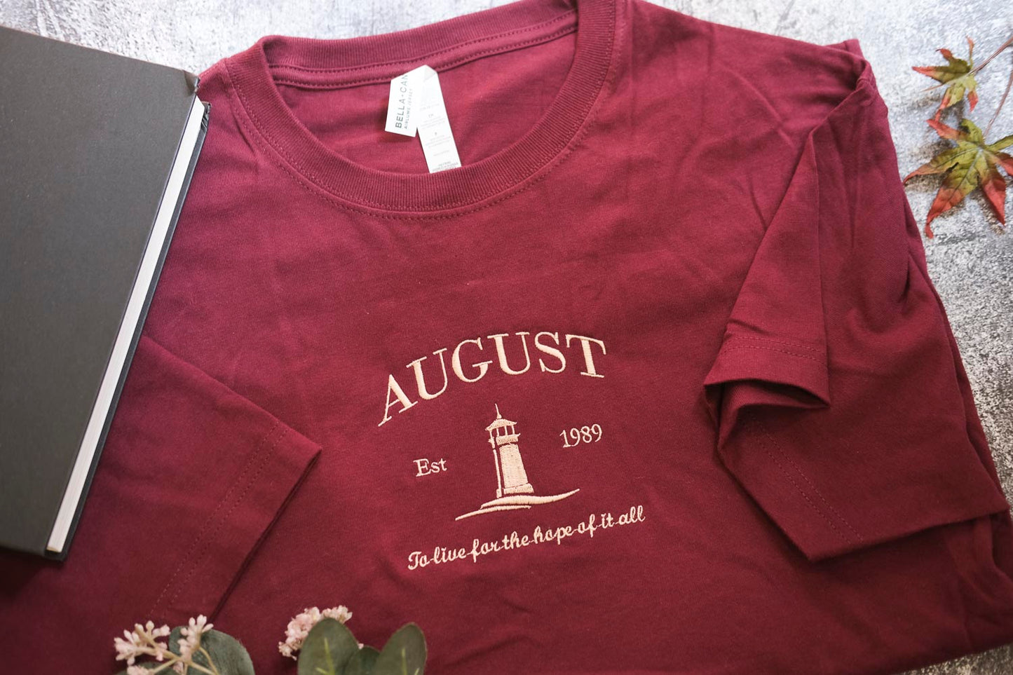 A burgundy t-shirt with the text "AUGUST Est. 1989" printed on it, along with a lighthouse graphic, is displayed on a surface next to a book and some leaves.