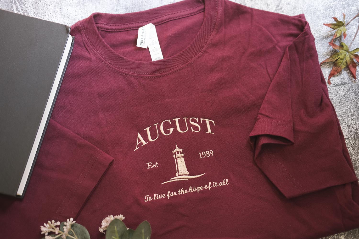 A burgundy t-shirt with the text "AUGUST Est. 1989" printed on it, along with a lighthouse graphic, is displayed on a surface next to a book and some leaves.