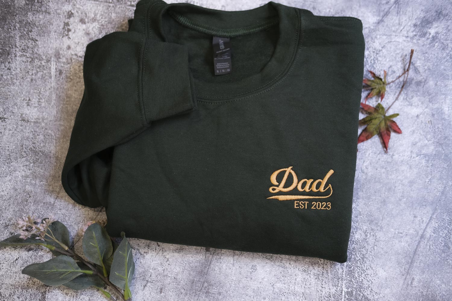 A dark green sweatshirt with the text "DAD EST 2023" printed on it, along with a small gold-colored logo.