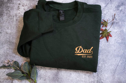 A dark green sweatshirt with the text "DAD EST 2023" printed on it, along with a small gold-colored logo.