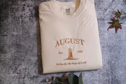 a white t-shirt with a printed design on it, placed on a textured gray surface. The design features a lighthouse and the text "AUGUST Est. 1989" along with the phrase "To live for the hope of it all".