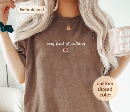 A woman with long blonde hair wearing a brown t-shirt with the text "very fond of walking" printed on it. She is holding a mug and has a necklace around her neck.
