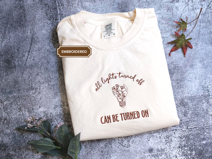 A white t-shirt with the text "all lights turned off" and an illustration of a light bulb is displayed on a gray surface, accompanied by some leaves.