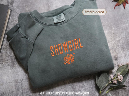 a gray sweatshirt with the text "SHOWGIRL" and a globe design printed on it, along with a label indicating that it is embroidered. The sweatshirt is placed on a gray surface, and there are some leaves and a book visible in the background.
