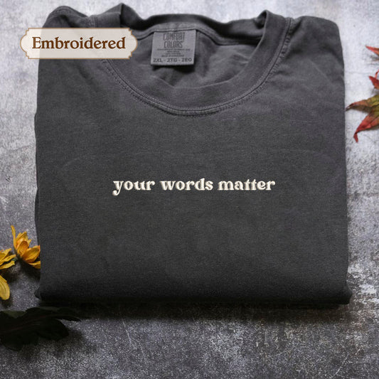 a gray t-shirt with the phrase "your words matter" embroidered on it, placed on a gray surface with some yellow flowers nearby.