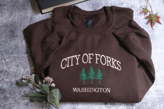 A brown sweatshirt with the text "CITY OF FORKS WASHINGTON" printed on it, along with a small green tree graphic, is laid out on a gray surface. There are also some leaves and a book visible in the background.