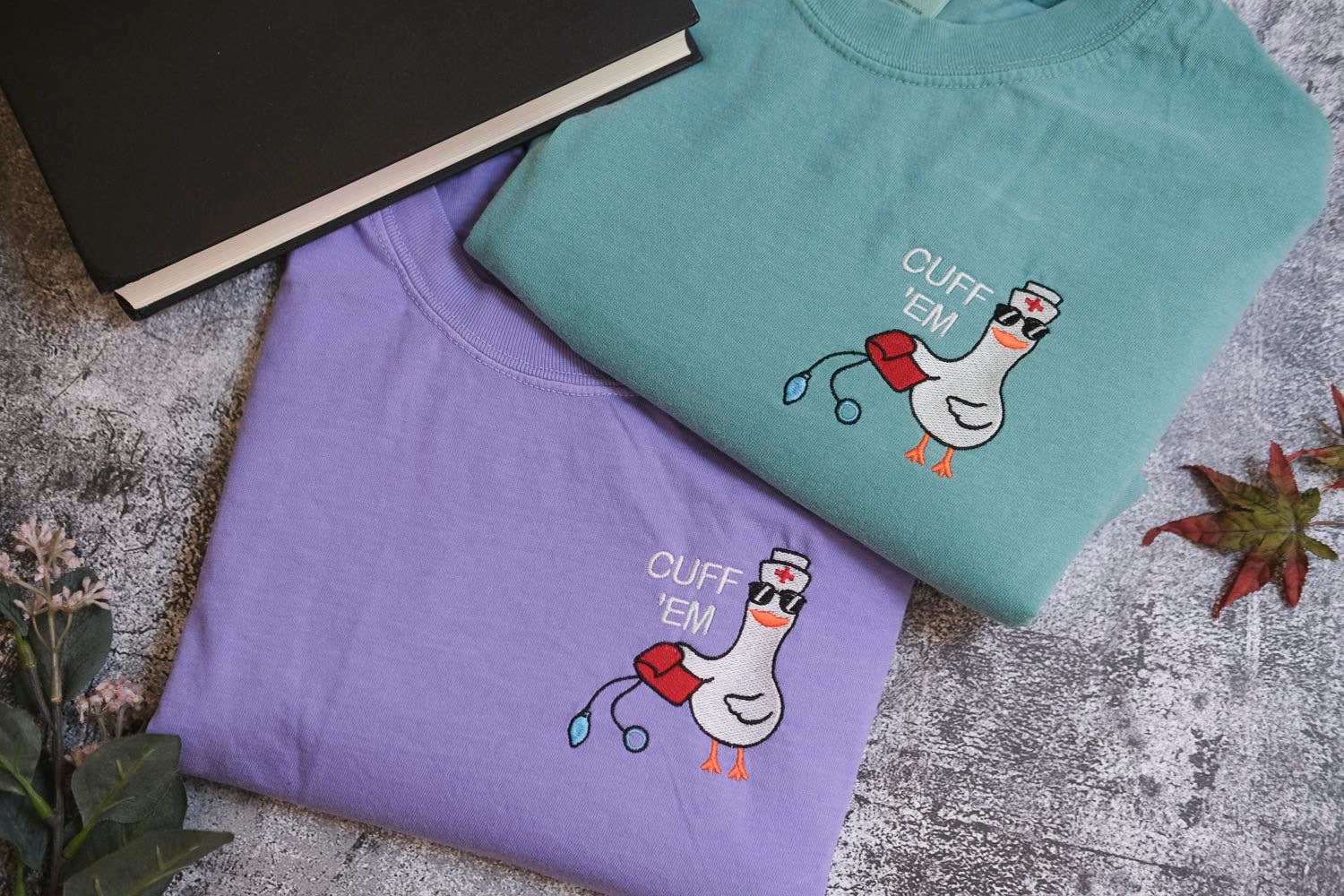 two t-shirts with a cartoon duck and the text "CUFF 'EM" printed on them, along with a black book and some leaves on the table.