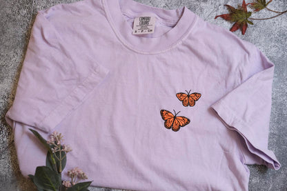 A light purple t-shirt with two orange butterflies printed on it, placed on a gray surface with some greenery and a small plant.