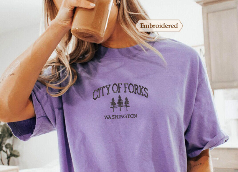 A person wearing a purple t-shirt with the text "CITY OF FORKS WASHINGTON" printed on it, holding a cup of coffee.
