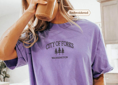 A person wearing a purple t-shirt with the text "CITY OF FORKS WASHINGTON" printed on it, holding a cup of coffee.