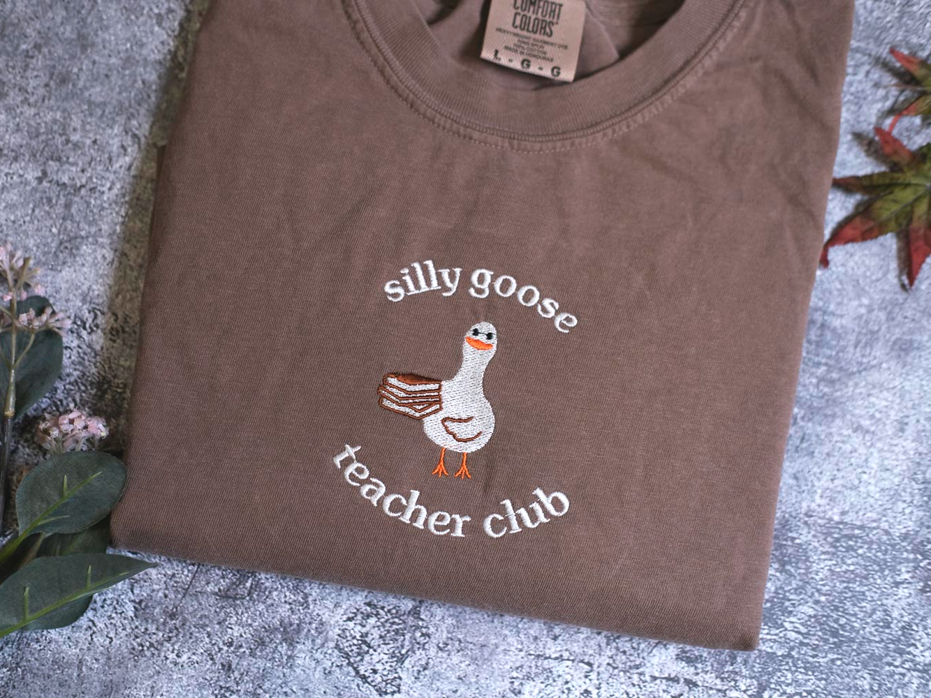 A brown t-shirt with a cartoon goose and the text "silly goose teacher chub" printed on it, along with a small label.