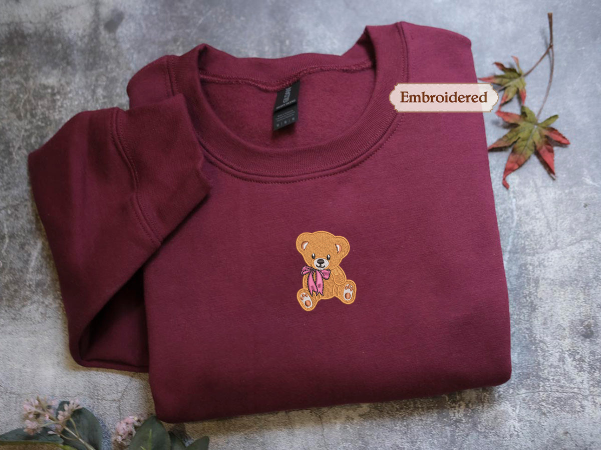 A burgundy sweatshirt with a teddy bear embroidered on the front, placed on a gray surface alongside some leaves.