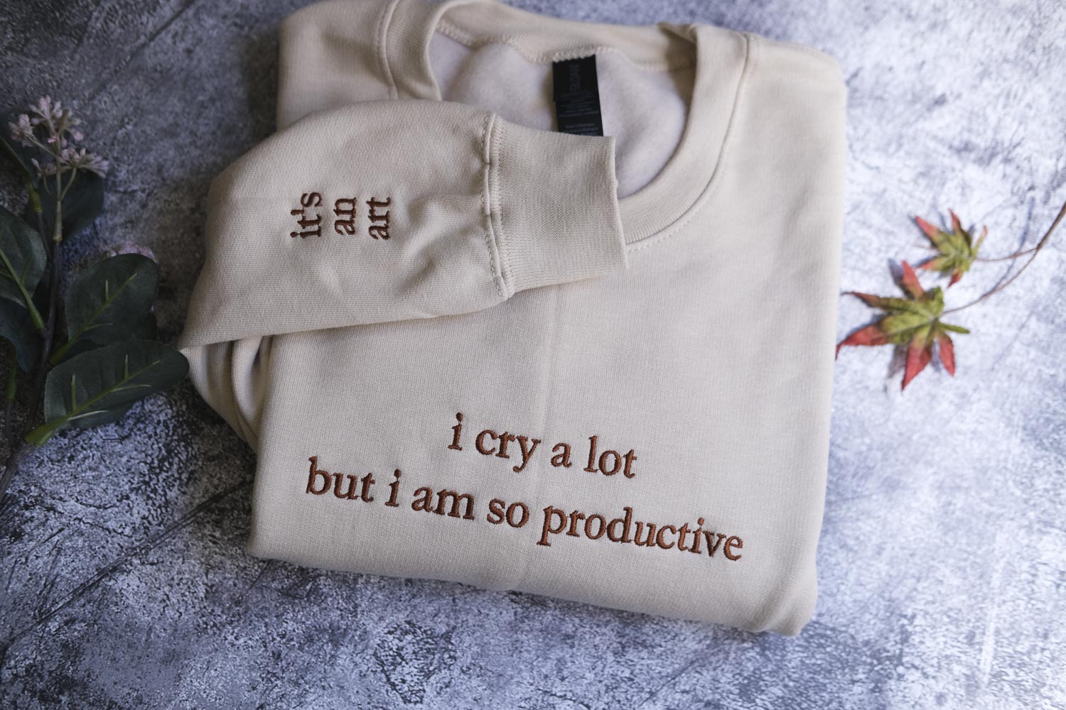 a beige sweater with the text "It's an art" and "I cry a lot but I am so productive" printed on it, along with a plant and a leaf in the background.