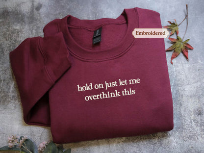 A burgundy sweatshirt with the text "hold on just let me overthink this" embroidered on it, along with a small embroidered label that says "Embroidered".