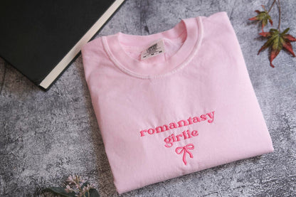 A pink t-shirt with the text "romantic girl" printed on it, along with a bow, is folded and placed on a gray surface next to a black book and some leaves.