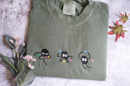 a green t-shirt with three small black creatures with white eyes and colorful accents, such as flowers, on it. The t-shirt is placed on a surface with some leaves and flowers, creating a natural and whimsical setting.
