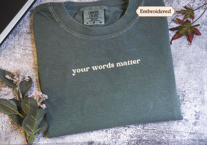 A gray t-shirt with the text "your words matter" printed on it, along with a small embroidered label. The t-shirt is placed on a surface with some leaves and flowers, and there is a small embroidered label visible.