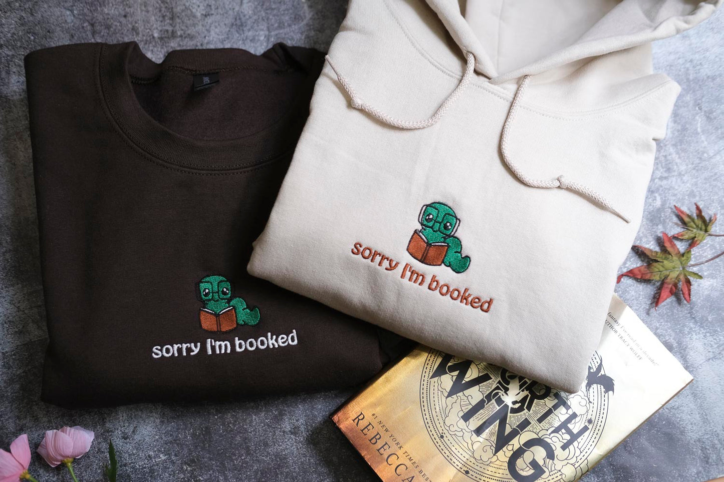 two hooded sweatshirts with a humorous text printed on them, along with a book and some flowers.