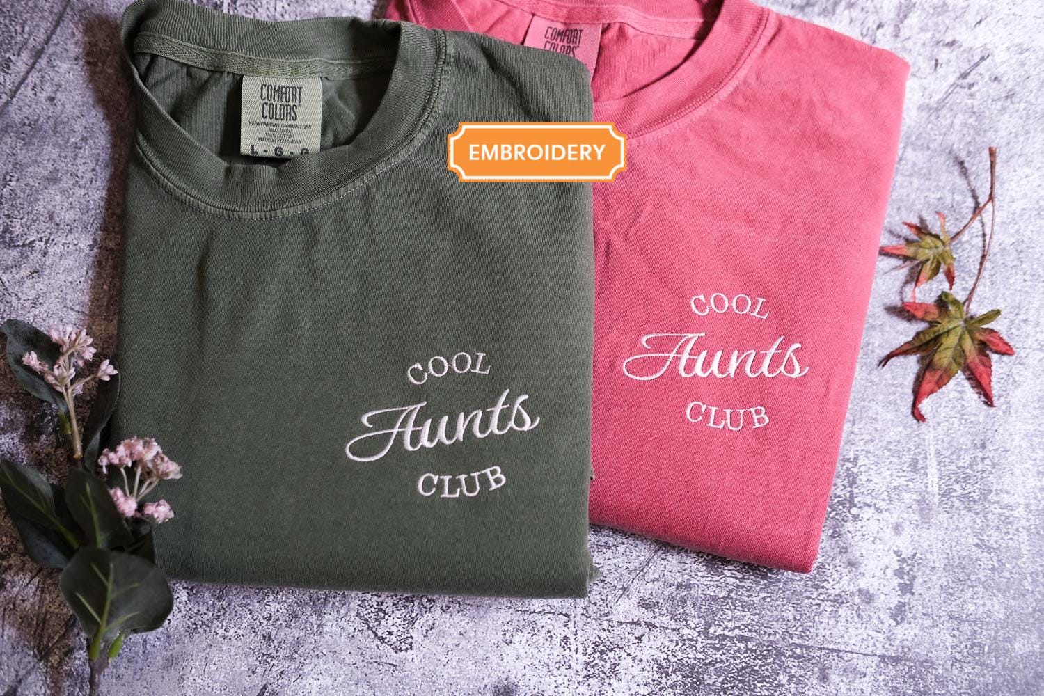 Two t-shirts are displayed on a gray surface, one in a dark green color and the other in a pink color. The dark green t-shirt has the text &quot;Cool Aunt&#39;s Club&quot; printed on it, while the pink t-shirt has the text &quot;Cool Aunt&#39;s Club&quot; printed on it. There are also some pink flowers and a fallen leaf in the background.